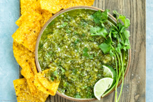 Overhead photo of Homemade Salsa Verde topped with lime wedges and fresh cilantro in a neutral colored with yellow tortilla chips scattered around the bowl and a chip inserted into the salsa.