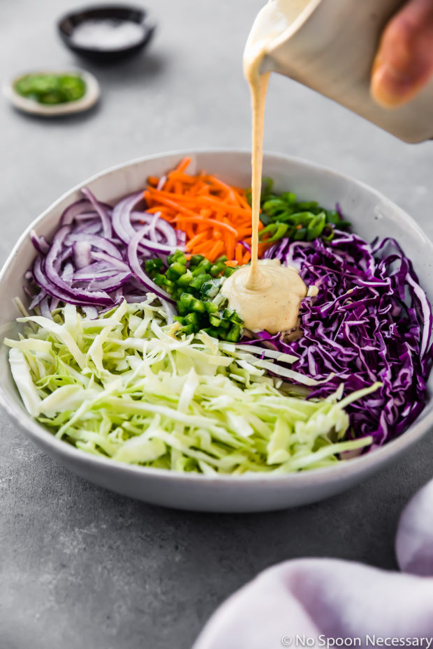 Angled shot of coleslaw sauce being poured into a bowl containing freshly shredded cabbage, carrots, jalapeños and red onions - step 3 in the recipe to make Spicy & Sweet Jalapeno Coleslaw.