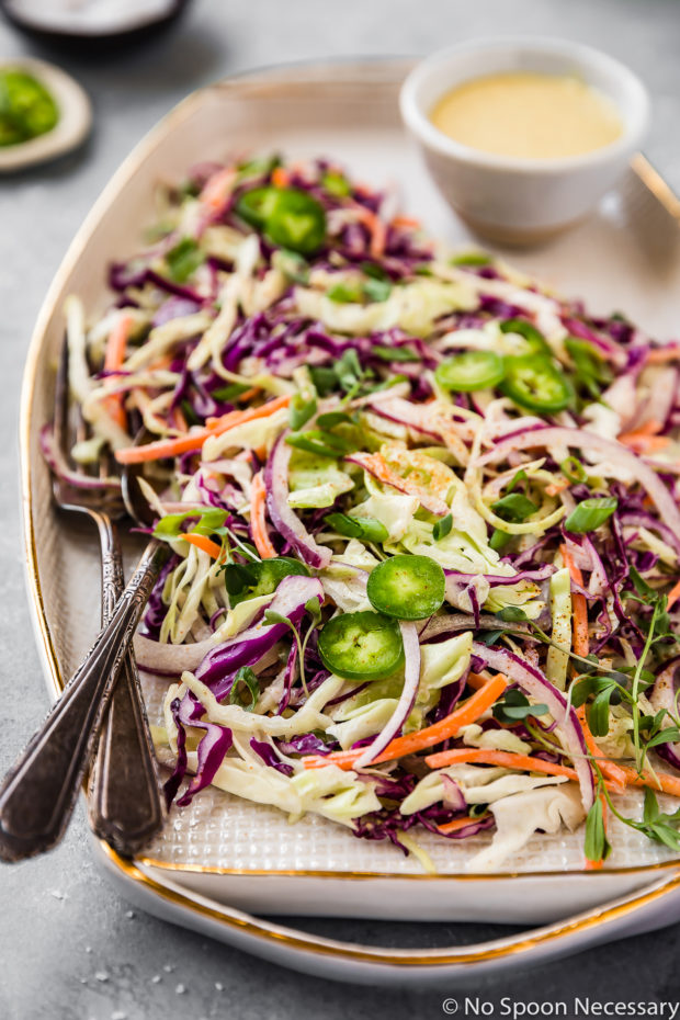 Angled shot of Spicy & Sweet Jalapeno Coleslaw on a white and gold trim platter with a fork and spoon tucked into the slaw and a ramekin of coleslaw sauce on the platter; with two small ramekins of salt and sliced jalapeños slightly blurred in the background.