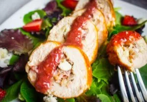 45 degree angle shot of a Chicken Roulade stuffed with Gorgonzola, Bacon & Caramelized Onions and drizzled with a Strawberry Balsamic Reduction on a plate of mixed greens.