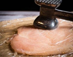 Angled shot of a meat mallet pounding out a chicken breast to make a chicken roulade.