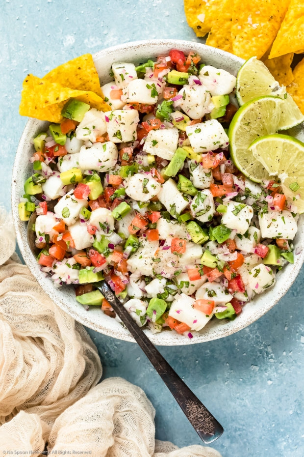 Overhead photo of Classic Fish Ceviche garnished with lime wedges in a white bowl with a serving spoon inserted into the ceviche and tortilla chips next to the bowl - photo of how to serve fish ceviche.