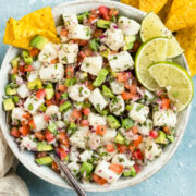 Overhead photo of Classic Fish Ceviche garnished with lime wedges in a white bowl with a serving spoon inserted into the ceviche and tortilla chips next to the bowl.