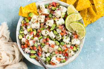 Overhead photo of Classic Fish Ceviche garnished with lime wedges in a white bowl with a serving spoon inserted into the ceviche and tortilla chips next to the bowl.