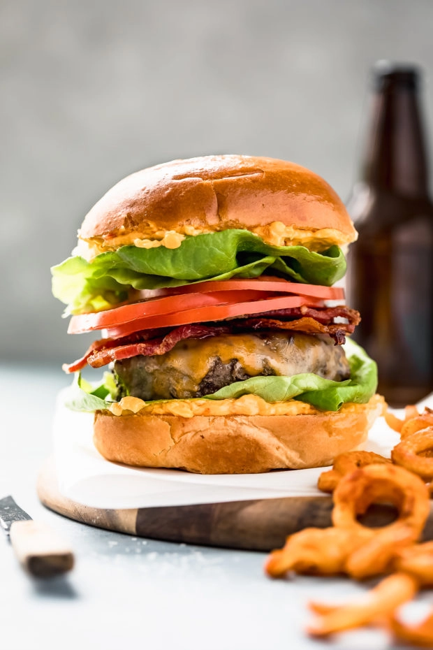 Straight on shot of a cheeseburger made with homemade ground beef and topped with bacon, sliced tomatoes and lettuce on a parchment paper lined wood serving tray with onion rings and a steak knife off to the side of the burger. 