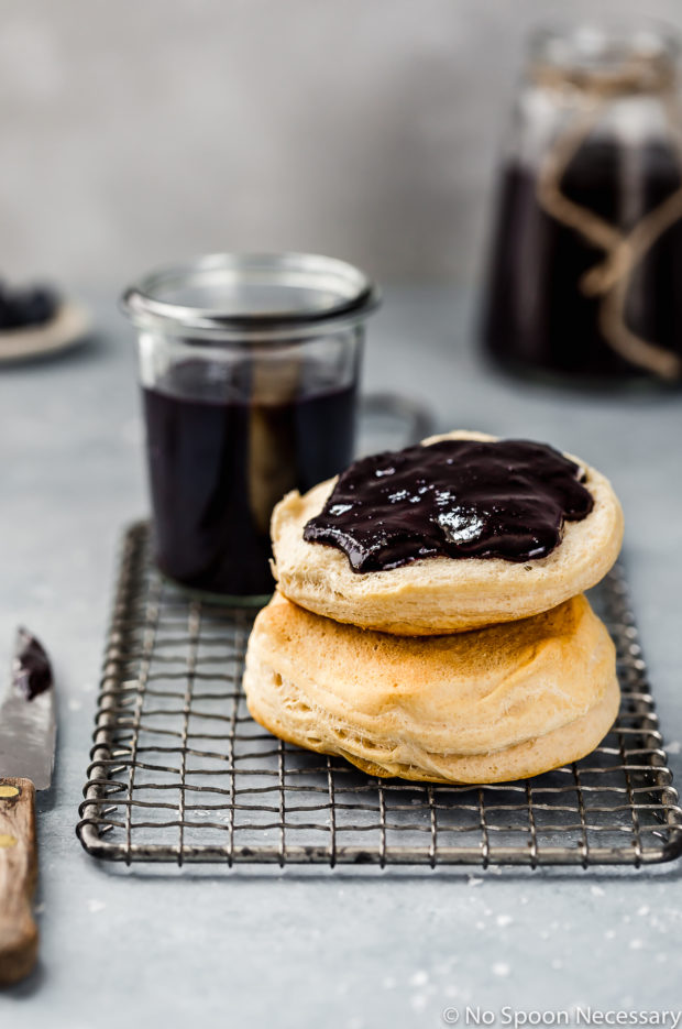 Straight on shot of a stack of biscuits smeared with Easy Slow Cooker Blueberry Butter on a small wire cooling rack, with two additional jars of homemade butter blurred in the background and a small knife off to the side.