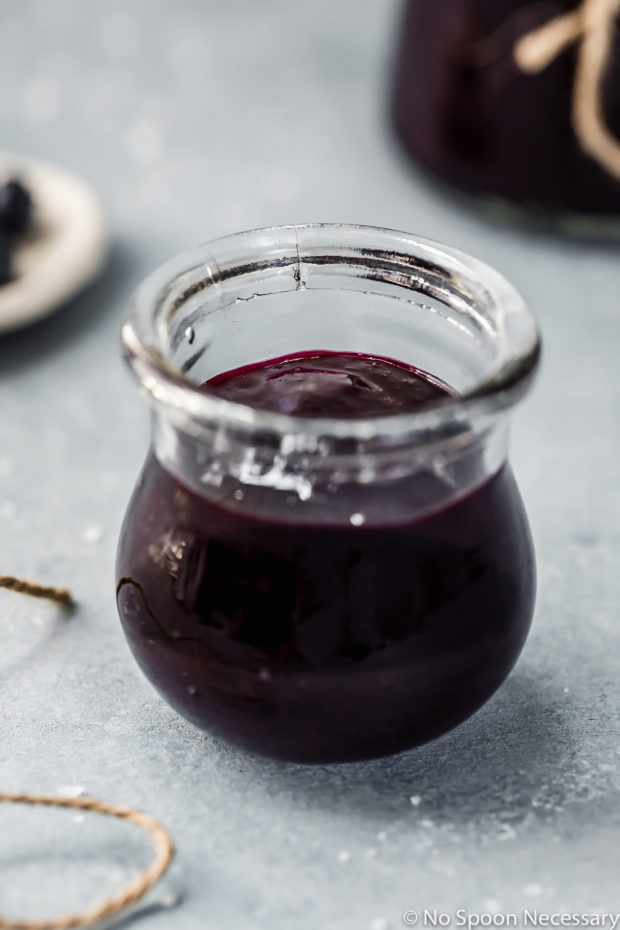 Angled shot of into a small jar of Easy Slow Cooker Blueberry Butter with a ramekin of fresh blueberries and an additional jar of homemade butter blurred and barely visible in the background. 