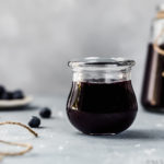 Straight on shot of a small jar of Easy Slow Cooker Blueberry Butter with an additional jar of homemade butter blurred in the background and fresh blueberries scattered around the jars.
