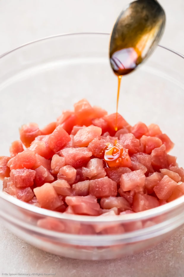 Angled photo of diced sashimi-grade tuna in a glass bowl with a spoon drizzling chili oil over the fish - action photo of step 1 of the spicy tuna recipe.