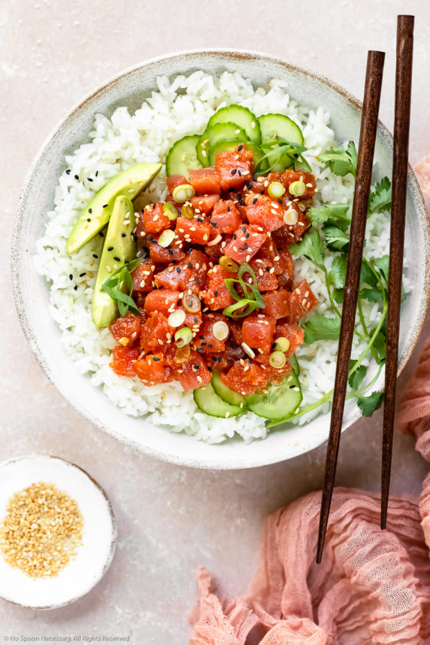 Overhead photo of Homemade Spicy Tuna Sushi Bowl garnished with slices of avocado, cucumber, scallions and fresh cilantro with a pair of wooden chopsticks resting on the bowl and a ramekin of sesame seeds and pink linen arranged around the bowl.