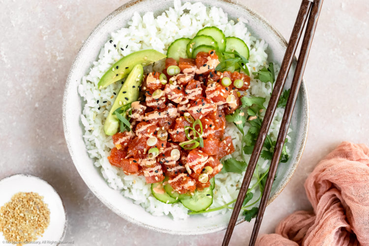 Overhead photo of Homemade Spicy Tuna Sushi Bowl garnished with spicy mayo, slices of avocado, cucumber, scallions and fresh cilantro with a pair of wooden chopsticks resting on the bowl and a ramekin of sesame seeds and pink linen arranged around the bowl.
