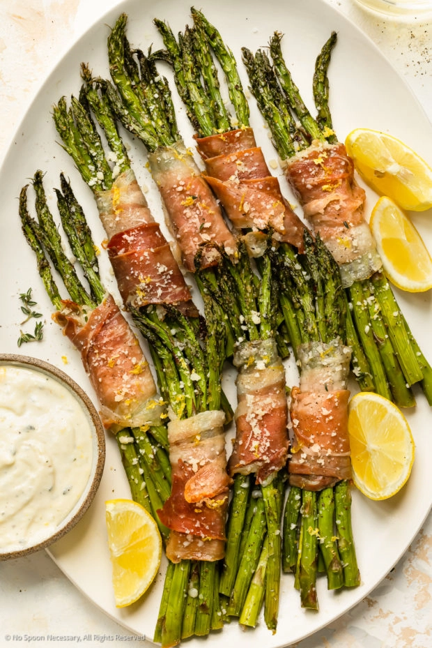 Overhead photo of asparagus prosciutto bundles with mayonnaise sauce for dipping on a white platter.