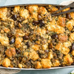 Overhead photo of baked Roasted Chestnut Stuffing in a blue baking dish with serving spoons inserted into the stuffing and glasses of wine and a pale neutral color linen next to the dish.
