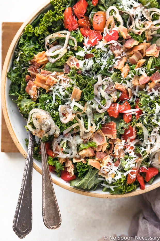 Overhead, up close shot of Warm Kale Salad topped with bacon, shallots, roasted red peppers and shredded parmesan cheese in a large serving bowl with serving spoons inserted into the salad.