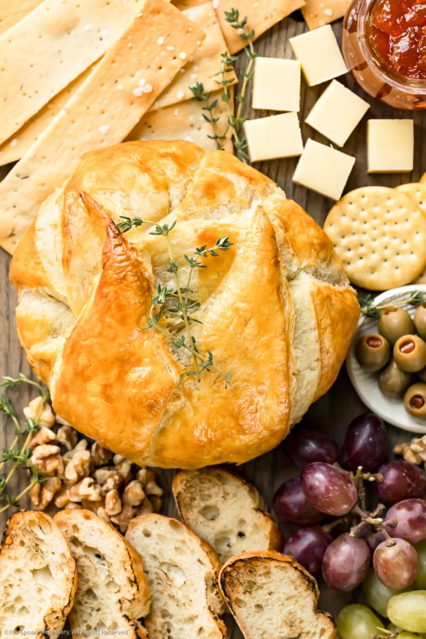 Overhead photo of freshly baked brie en croute garnished with fresh thyme leaves on a cheeseboard with crackers, slices of bread, grapes, olives and nuts.