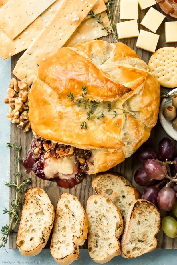 Overhead photo of Baked Brie en Croute that has been cut into on a large cheese board surrounded by crackers, slices of bread, olives, jam, grapes and nuts.