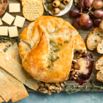 Overhead photo of Baked Brie en Croute on a large cheese board surrounded by crackers, slices of bread, olives, jam, grapes and nuts.