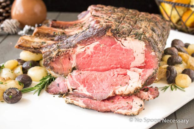 Slightly angled shot of a Herb Crusted Standing Rib Roast on a large white platter with roasted mushrooms and onions.