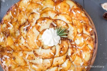 Overhead shot of a Asiago, Roasted Garlic & Rosemary Potato Galette topped with a dollop of sour cream and rosemary sprig on a gray wood surface.