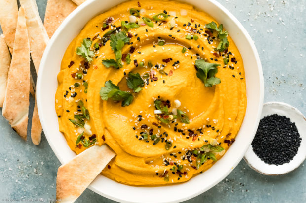Overhead photo of creamy carrot dip garnished with fresh cilantro and sesame seeds in a white serving bowl with a slice of pita bread inserted into the dip and more slices of pita bread next to the bowl.