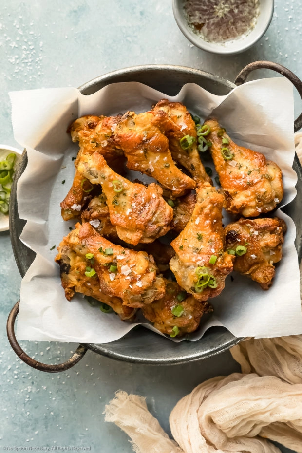 Overhead photo of Salt & Vinegar Chicken Wings in a metal serving bowl lined with parchment paper with ramekins of dipping sauce and sliced scallions arranged around the bowl.