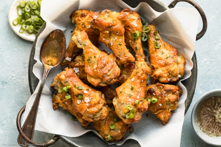 Overhead, landscape photo of Salt & Vinegar Chicken Wings in a metal serving bowl lined with parchment paper with ramekins of dipping sauce and sliced scallions arranged around the bowl.