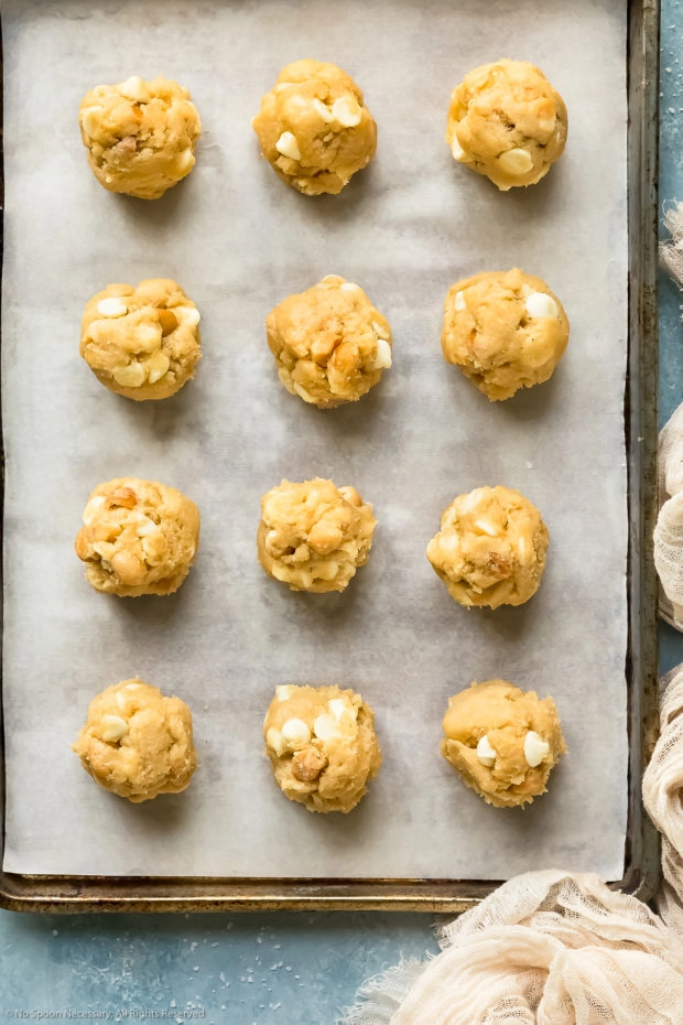 Overhead photo of white chocolate macadamia nut cookie dough rolled into individual balls on a parchment paper lined cookie sheet.