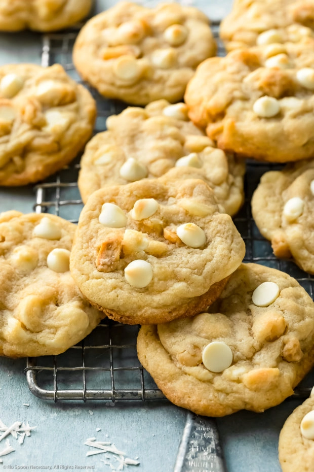 Angled photo of White Chocolate Macadamia Nut Cookies scattered on top of each other on a wire rack on a blue surface.