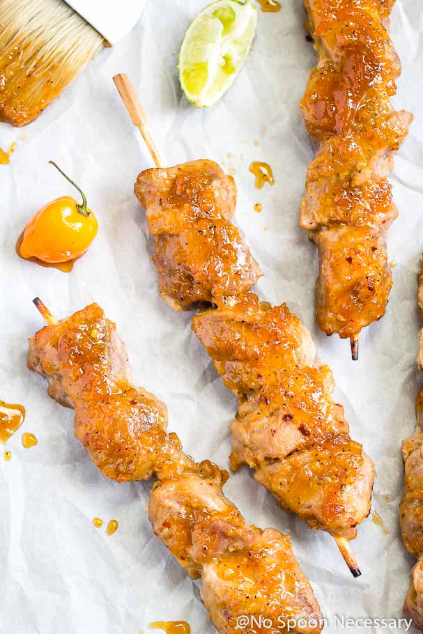 Overhead shot of three Apricot, Habanero & Honey Glazed Pork Tenderloin Skewers on a piece of crinkled parchment paper with a culinary brush, habanero and lime wedge.