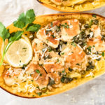 Overhead shot of Healthy Chicken Piccata Spaghetti Squash Boats garnished with a slice of lemon and sprig of fresh parsley on a parchment paper lined wood serving board with a ramekin of chopped parsley next to the board.