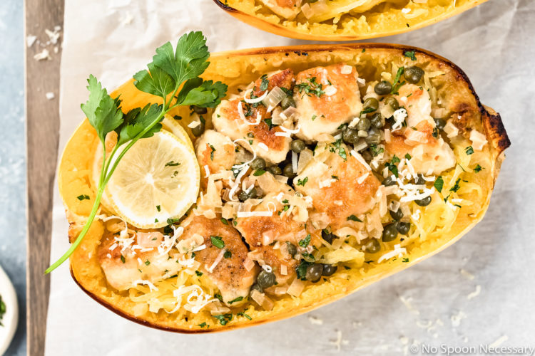 Overhead shot of Healthy Chicken Piccata Spaghetti Squash Boats garnished with a slice of lemon and sprig of fresh parsley on a parchment paper lined wood serving board with a ramekin of chopped parsley next to the board.