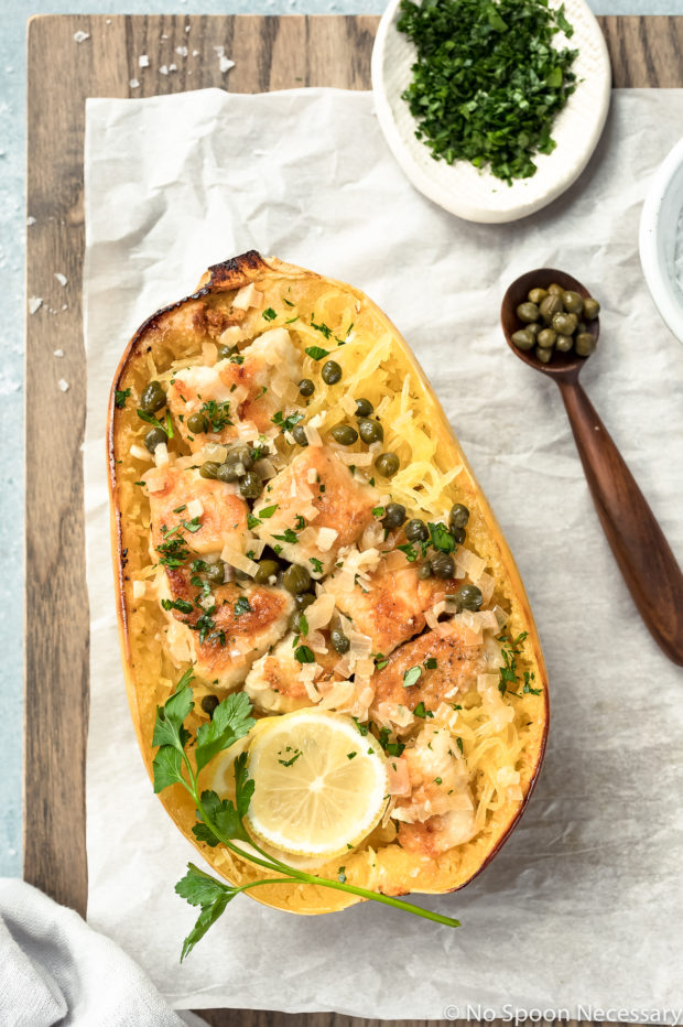 Overhead shot of Healthy Chicken Piccata Spaghetti Squash Boats garnished with a slice of lemon and sprig of fresh parsley on a parchment paper lined wood serving board with a ramekin of chopped parsley and spoon full of capers next to the squash.
