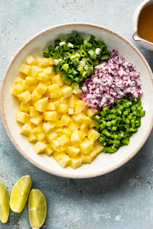 Overhead photo of all the ingredients needed to make tropical pineapple salsa for salmon in a large white bowl with fresh lime wedges next to the bowl. 