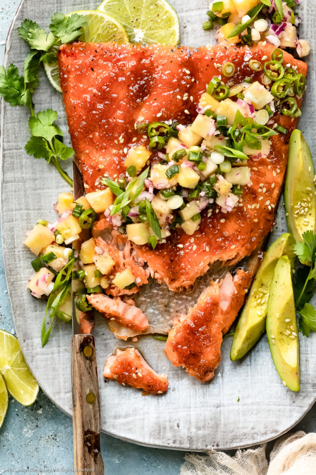 Overhead photo of Baked Honey Sriracha Salmon topped with tropical pineapple salsa on a gray serving platter with a portion of the salmon flaked off to showcase how tender the interior is.