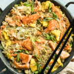 Overhead, landscape photo of Salmon Quinoa Fried Rice in a metal wok with a pair of chopsticks resting on the side of the wok and a ramekin of sesame seeds and pale tan napkin next to the wok.
