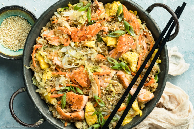 Overhead, landscape photo of Salmon Quinoa Fried Rice in a metal wok with a pair of chopsticks resting on the side of the wok and a ramekin of sesame seeds and pale tan napkin next to the wok.
