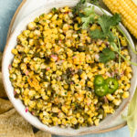 Overhead, landscape photo of Roasted Sweet Corn Salsa garnished with fresh cilantro in a large white bowl with an ear of corn and tortilla chips next to the bowl.