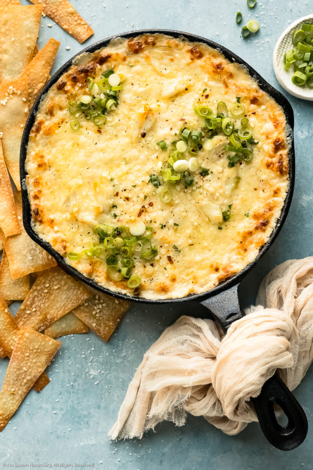 Overhead photo of Baked Crab Rangoon Dip garnished with fresh scallions in a black cast iron skillet with wonton chips and a ramekin of sliced scallions next to the skillet.