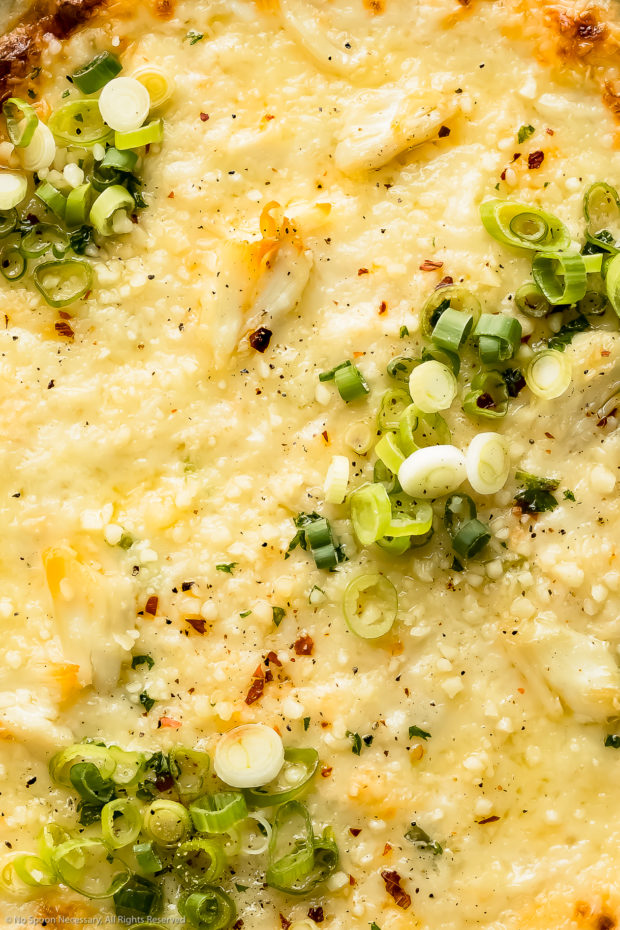 Overhead, extreme close-up photo of crab dip garnished with crushed red pepper flakes and sliced scallions.