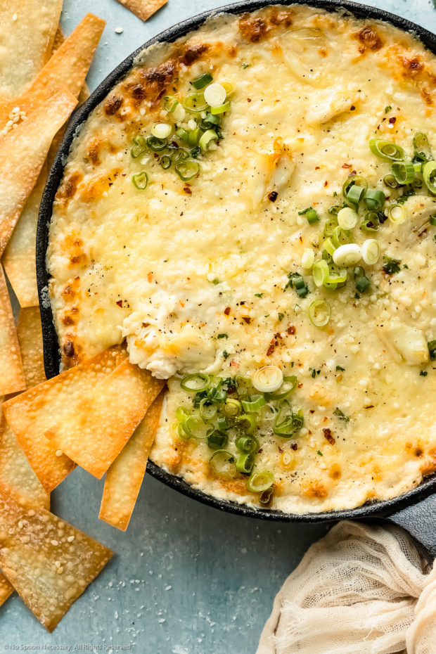 Overhead photo of Baked Crab Rangoon Dip garnished with fresh scallions in a cast iron skillet with a few wonton chips inserted into the dip and more chips scattered next to the cast iron skillet.