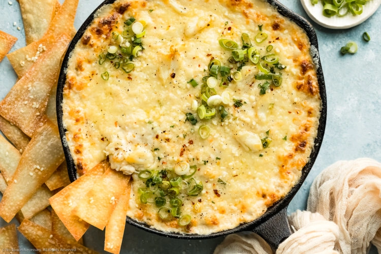 Overhead photo of Hot Crab Rangoon Dip garnished with fresh scallions in a cast iron skillet with a couple wonton chips inserted into the dip and more chips plus a ramekin of sliced scallions next to the skillet.