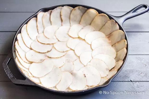Angled photo of thin slices of potato lining the inside of a cast iron skillet. 