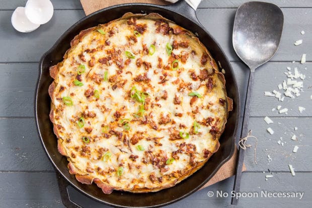 Overhead shot of Spanish Potato Crusted Frittata in a cast iron skillet on top of a wood board with a large serving spoon, grated cheese and egg shells surrounding the skillet.