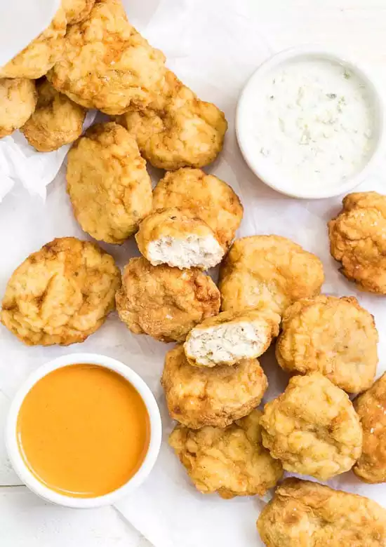 Overhead shot of a pile of Copycat McDonald's Buffalo Chicken Nuggets on a piece of parchment paper with small ramekins of buffalo sauce and blue cheese; with a nugget torn in half in the middle of the pile