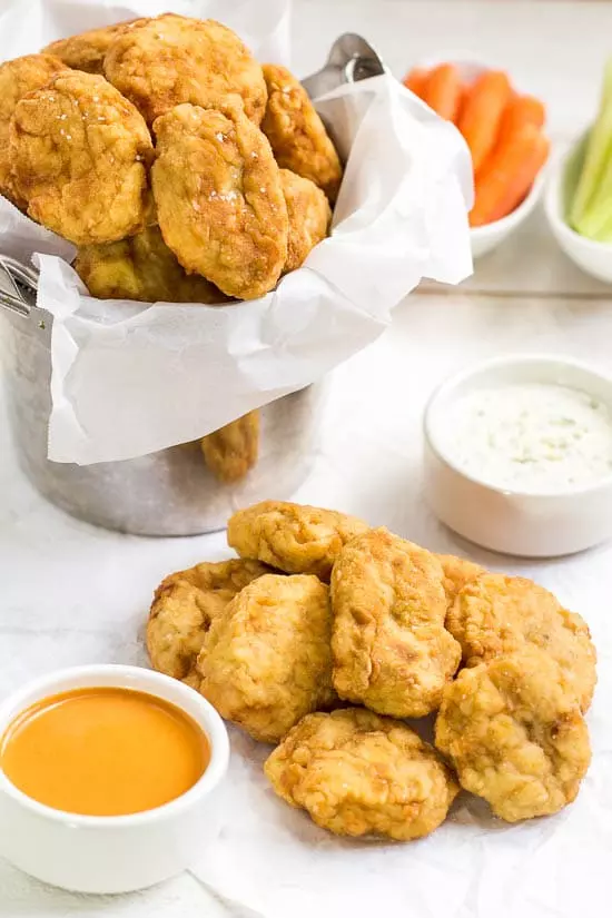 45 degree angle shot of a pile of Copycat McDonald's Buffalo Chicken Nuggets on a piece of parchment paper with a small ramekins of buffalo sauce, blue cheese and a large bucket of more nuggets surrounding. 