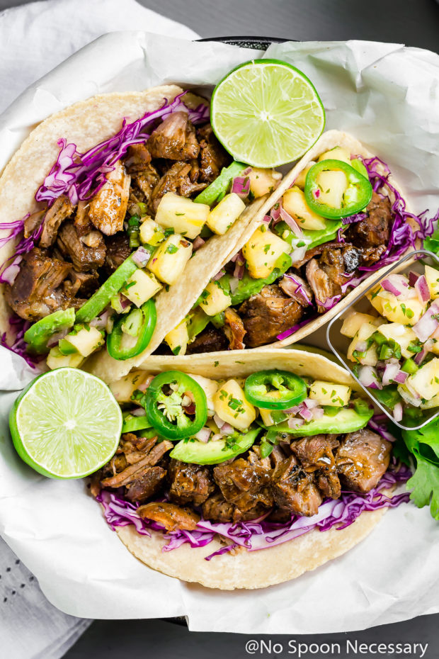 Overhead shot of a bowl with parchment paper topped with Jerk Carnitas Tacos with Pineapple Jalapeno Salsa and lime wedges; with a pale purple linen underneath the bowl.