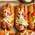 Overhead photo of four bacon wrapped hot dogs topped with lettuce, tomatoes, onions and mayonnaise.
