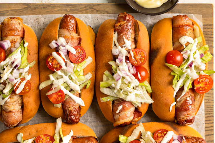Overhead photo of four bacon wrapped hot dogs topped with lettuce, tomatoes, onions and mayonnaise.