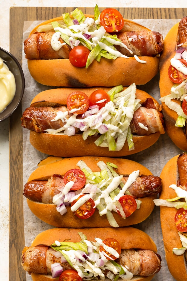 Overhead photo of four oven baked bacon wrapped hot dogs topped with shredded lettuce, cherry tomatoes and mayonnaise sauce.