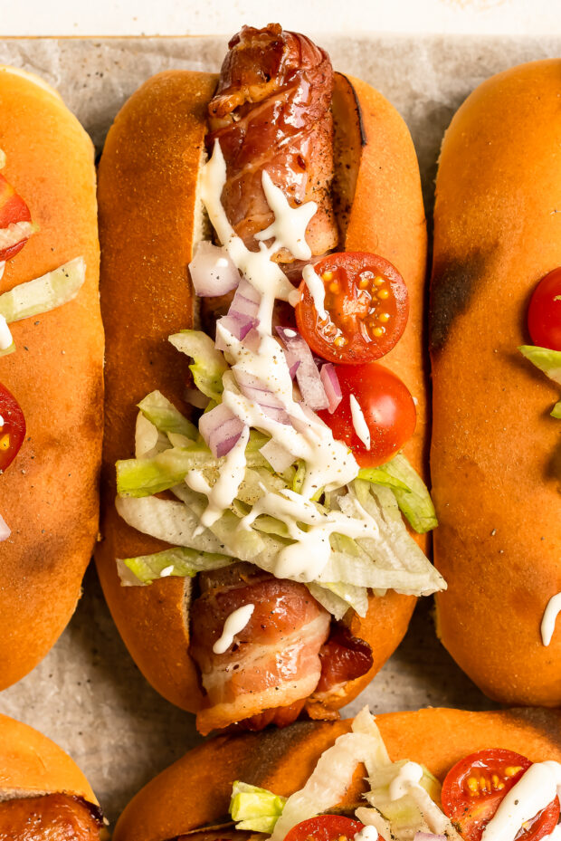 Overhead, close up photo of a bacon hot dog topped with lettuce, tomatoes and red onions in a toasted bun.
